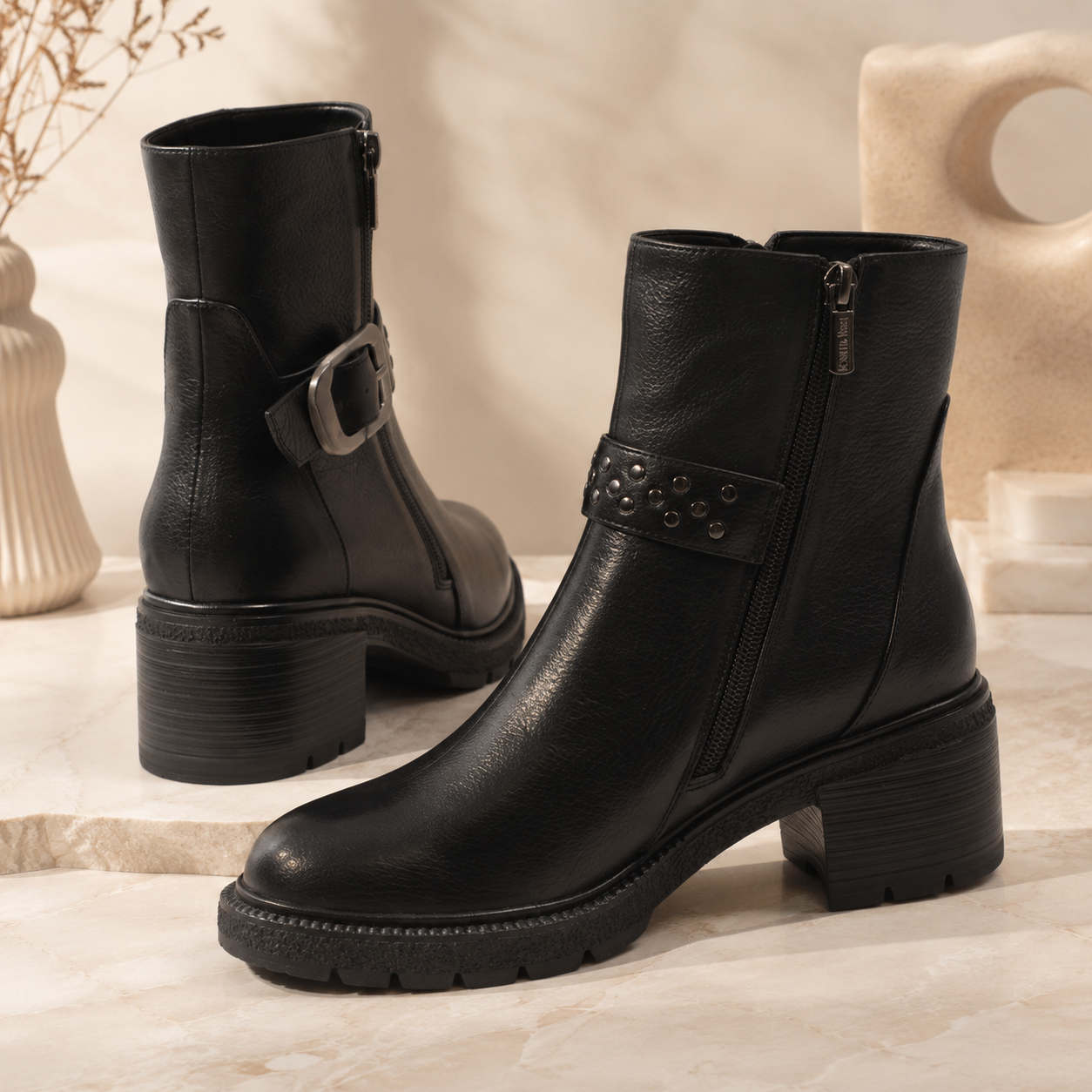 A pair of black leather ankle boots with chunky block heels, side zippers, and decorative buckle and stud details, displayed on a light marble surface with neutral decorative props in the background.
