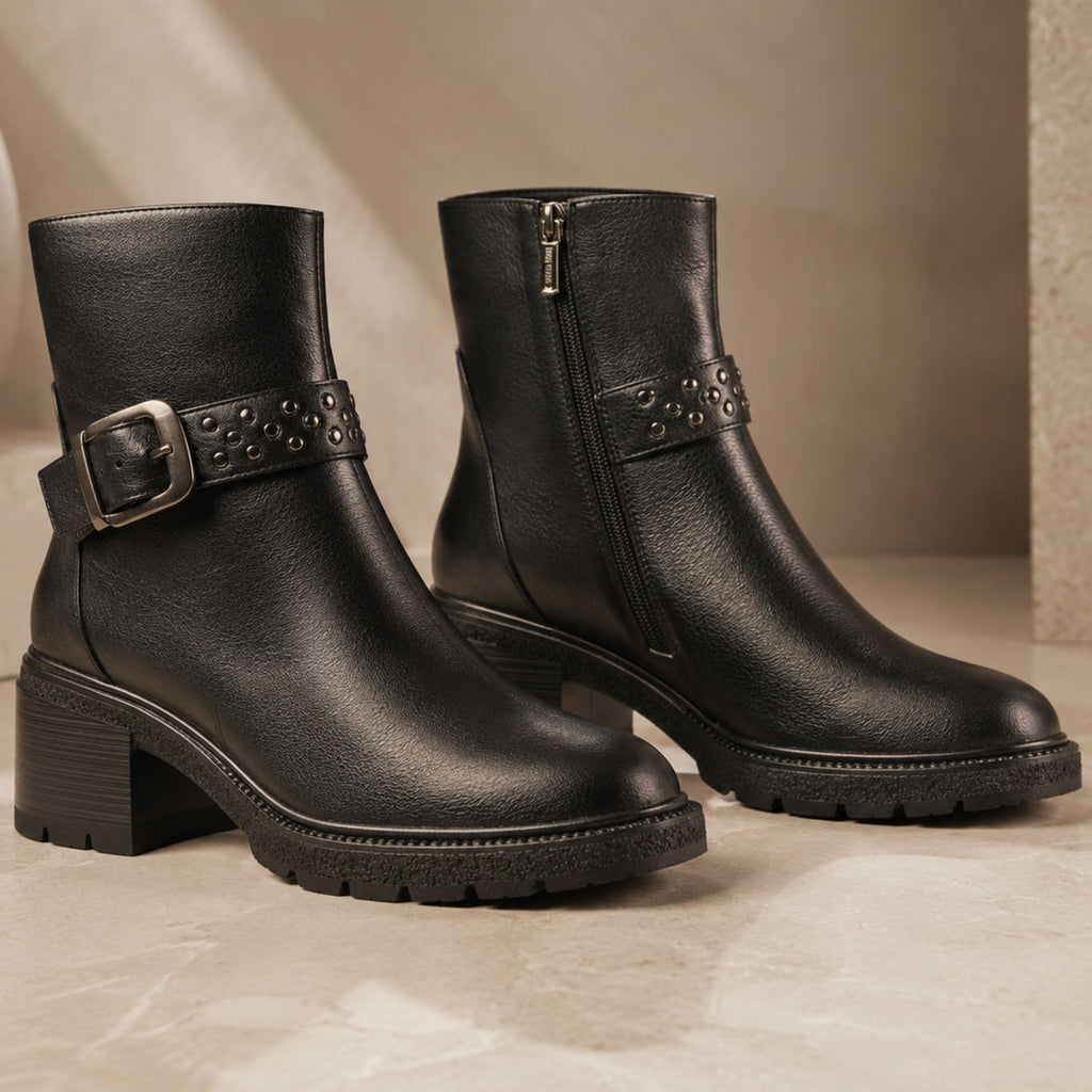 A pair of black leather ankle boots with chunky lug soles, a side zipper, and a studded strap with a silver buckle on the outer side, displayed on a beige stone surface.