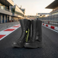 A pair of black Trifecta motorcycle boots with bright yellow-green zipper accents, displayed on a racetrack asphalt surface with grandstands in the background.