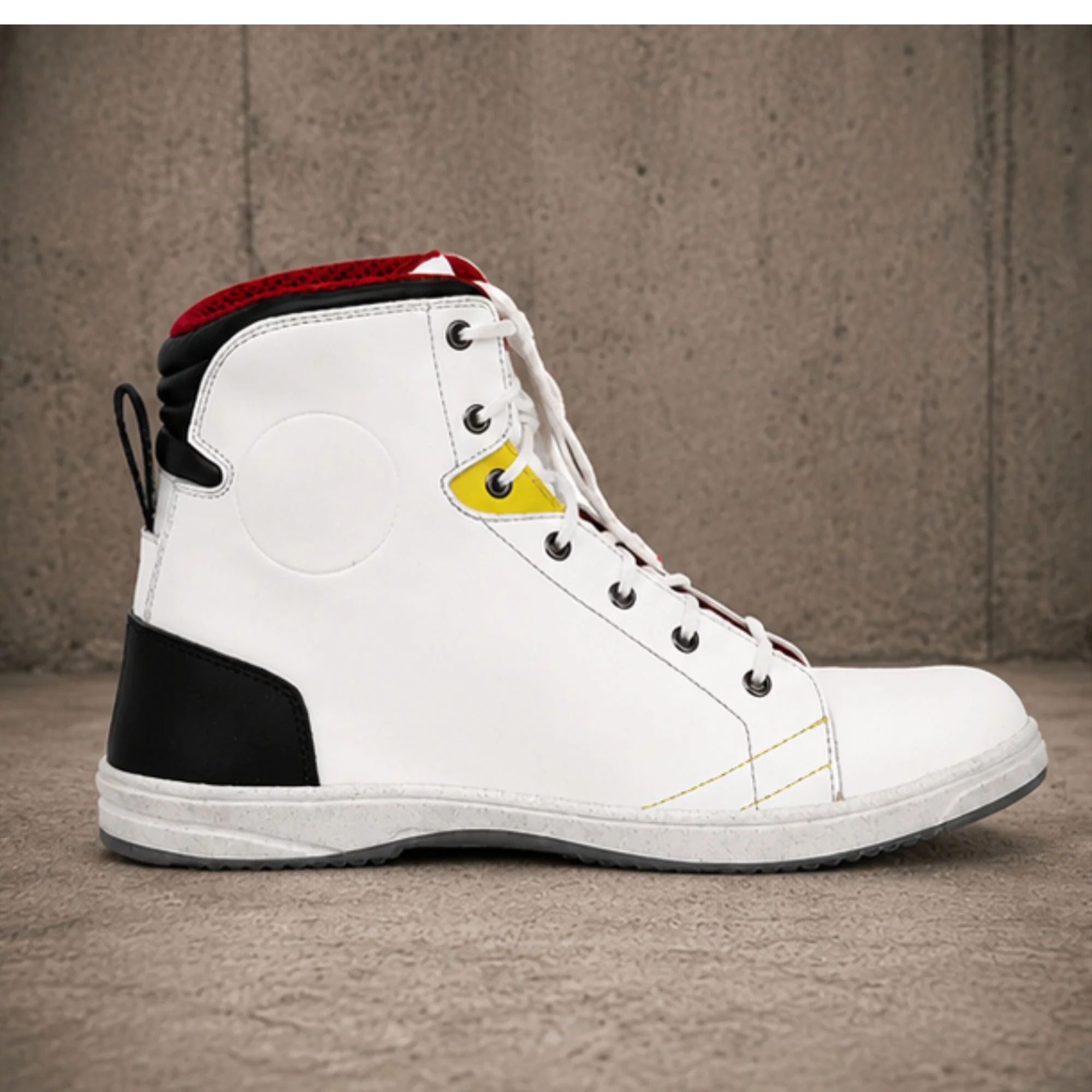A high-top white leather sneaker with black and red accents, featuring a yellow decorative tab near the lace area, photographed against a textured concrete wall and floor.